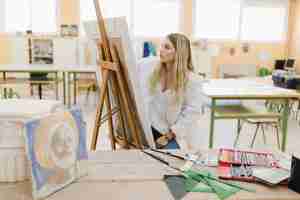 Foto gratuita giovane donna bionda che si siede nella pittura dell'officina sul cavalletto