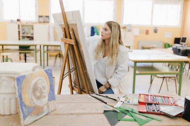 Блондинка молодая женщина, сидя в мастерской, живопись на мольберте