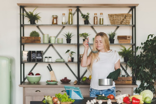 Blonde young woman smelling soup from the ladle in the kitchen