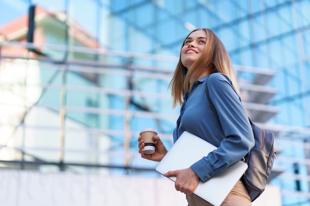 Blonde young woman smiling portrait holding laptop and coffee, wearing blue gentle shirt over modern building