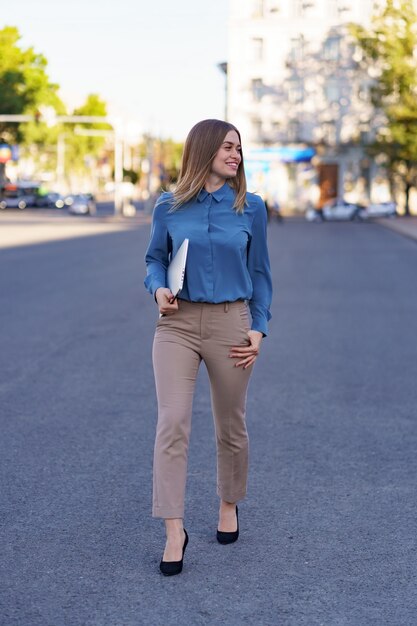 Blonde young woman smiling portrait wearing blue gentle shirt holding a laptop while walking