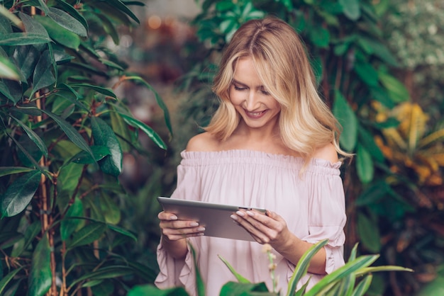 デジタルタブレットを見て緑の植物の近くに立っている金髪の若い女性