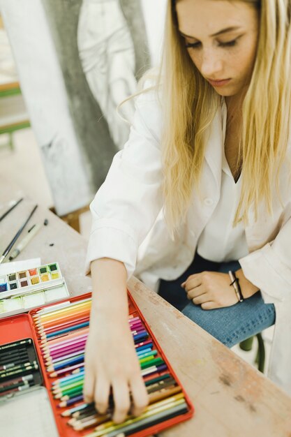 Blonde young woman taking colored pencil from case on wooden table