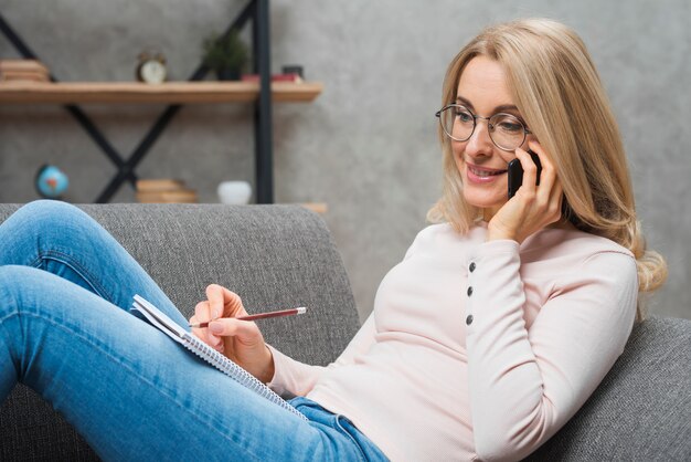 鉛筆でスパイラルメモ帳に書いて携帯電話で話している金髪の若い女性