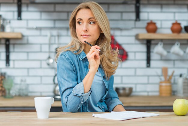 Blonde young woman thinking while writing on notebook over the wooden table