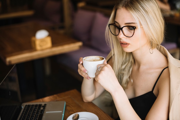 カフェでコーヒーカップを保持している眼鏡を着ているブロンドの若い女性