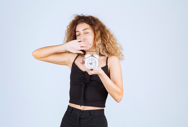 Blondie girl with an alarm clock looks sleepy. 