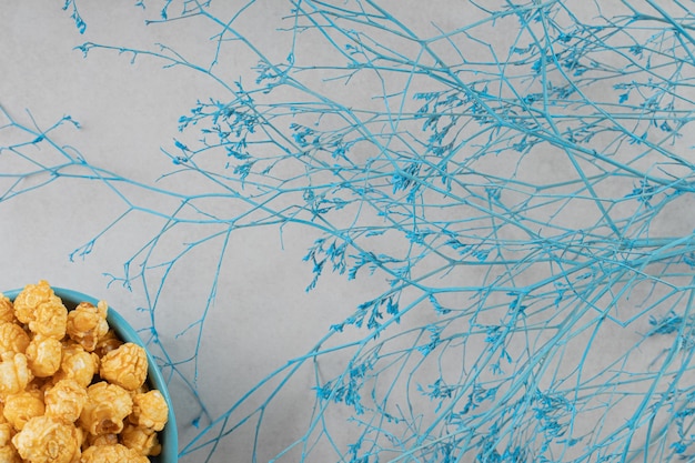 Blue bowl of caramel flavored popcorn next to decorative branches on marble background. 