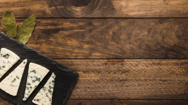 Blue cheese slice arrange on black slate with bay leaves over old wooden desk