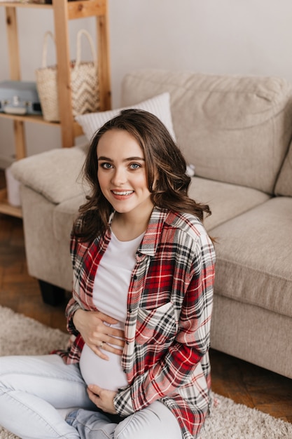 Blue-eyed brunette pregnant woman in plaid shirt and white tee gently touches belly and looks into front