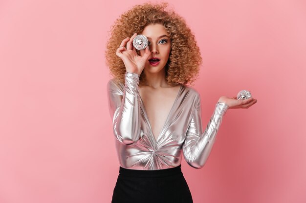 Blue-eyed curly blonde woman dressed in shiny top looking at camera and holding small disco balls on pink space.
