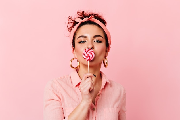 Blue-eyed curly girl in pink headband and massive gold earrings eating candy on pink space.