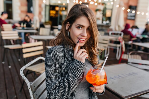 屋外カフェでカクテルを飲む灰色のコートの青い目の女性モデル