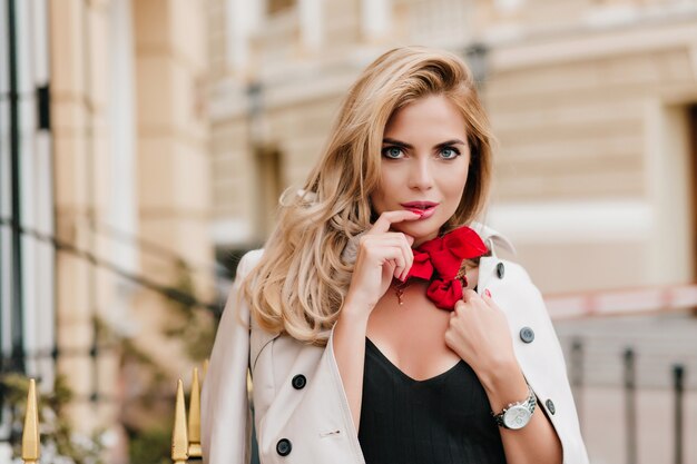 Blue-eyed female model in trendy red scarf posing with pleasure in new outfit standing beside building