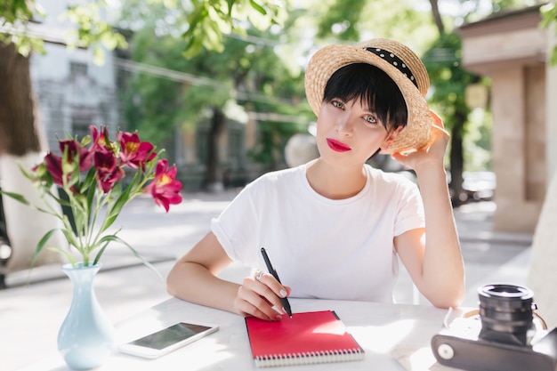 Blue-eyed female student in straw hat doing homework in outdoor cafe, sitting with pen and notebook