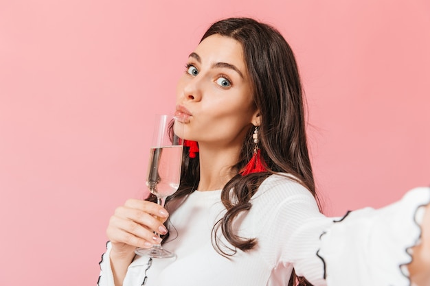 La ragazza dagli occhi azzurri fa selfie su sfondo rosa con un bicchiere di spumante.