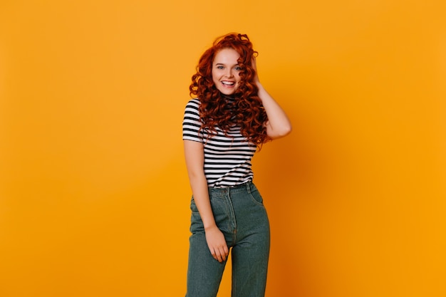 Blue-eyed girl with red curly hair smiling and looking into camera. Portrait of young woman in denim pants and black-white T-shirt.