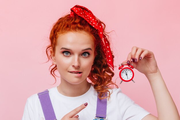 Blue-eyed girl with red ribbon in her hair bites her lip and points to alarm clock