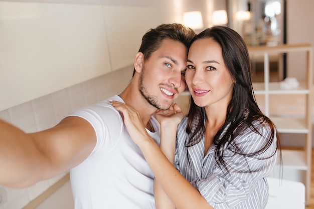 Blue-eyed man in white t-shirt making selfie with lovely black-haired woman in home