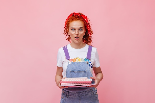 Blue-eyed red-haired girl in jeans outfit and glasses looks at front and holds pink books on isolated wall