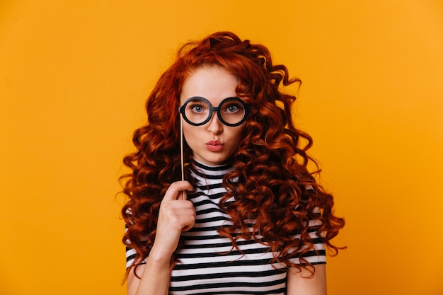 Blue-eyed red-haired woman in striped T-shirt blows kiss and holds toy glasses on stick, posing against orange space.