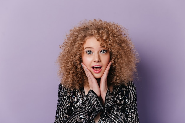 Blue-eyed woman in amazement posing against lilac space. Portrait of curly blonde in top with silver sequins.