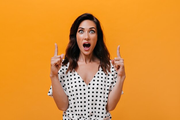 Blue-eyed woman looks at camera in surprise and points her finger up to place for text