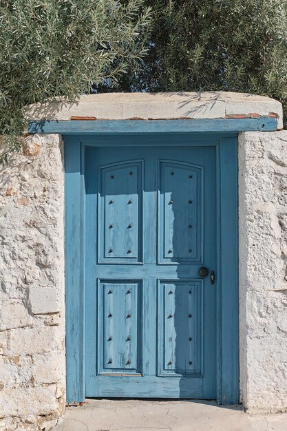 집의 돌 흰 벽에 있는 파란색 오래된 문 올리브 가지가 문 세로 프레임 지중해 해안 골동품 빈티지 스타일 그리스 여행 시간 배경 아이디어 위에 걸려 있습니다.
