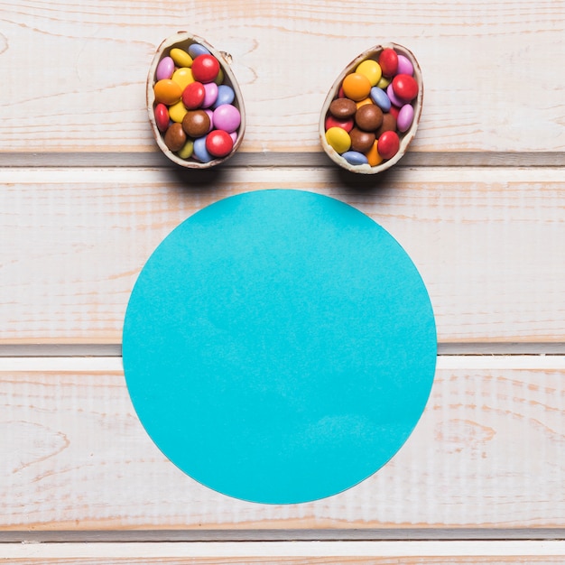 Free photo blue paper circular frame with easter eggs filled with colorful gem candies over the wooden desk