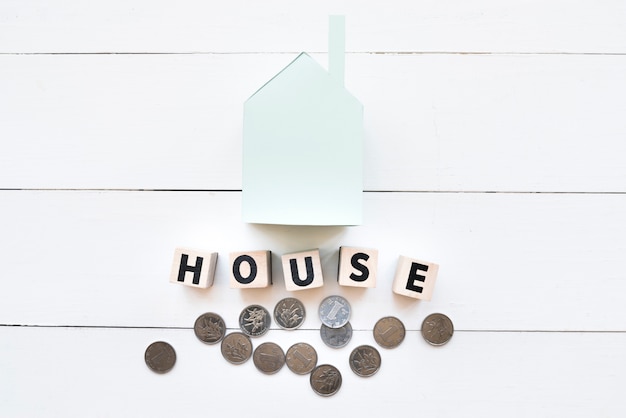 Blue paper house model with wooden blocks and coins on white wooden backdrop