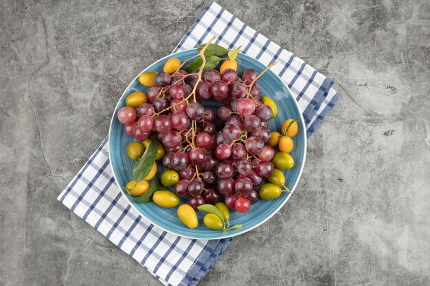 Blue plate of kumquat fruits and red grapes on marble surface. 