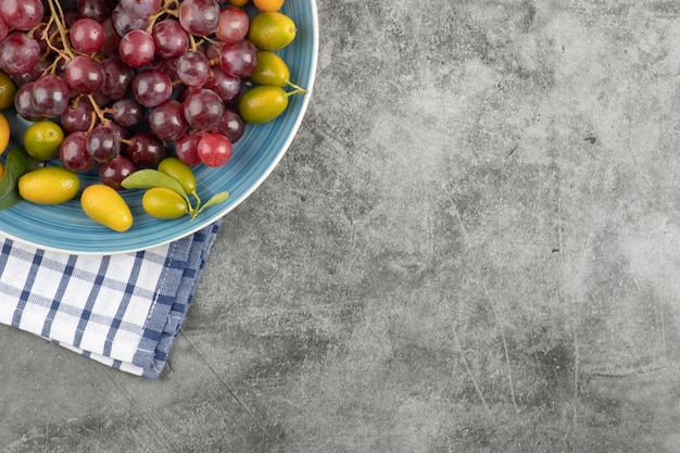 Blue plate of kumquat fruits and red grapes on marble surface.