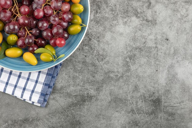 Blue plate of kumquat fruits and red grapes on marble surface.