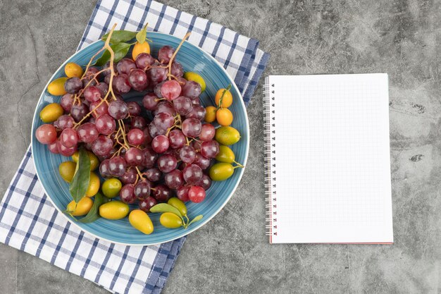 Blue plate of kumquat fruits and red grapes with empty notebook. 