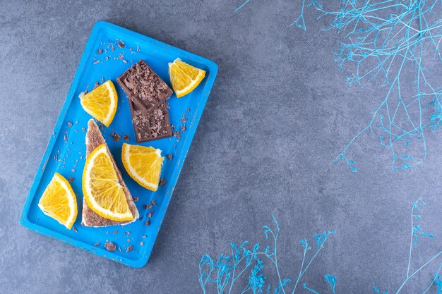 装飾的な枝の横にある青い大皿、大理石の表面にケーキスライス、チョコレートプレート、オレンジスライスがあります