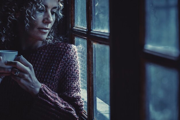夜にお茶を飲んで家の窓の外を見ている青い色調の孤独な女性の肖像画。悲しみと不眠症の概念。魅力的な大人の女性だけのコンセプト Premium写真