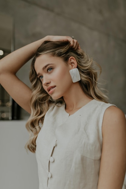 Blueeyed blonde curly woman touches hair and demonstrates her big beautiful white earrings attrac