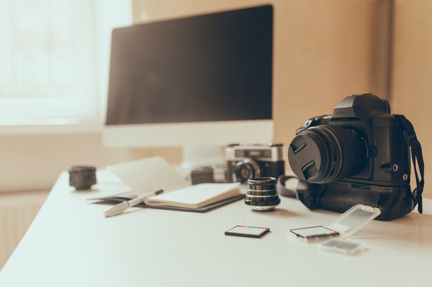 Blur photo of workplace with camera and memory cards on foreground. Modern computer stands on white table with notebook and pen lying beside.