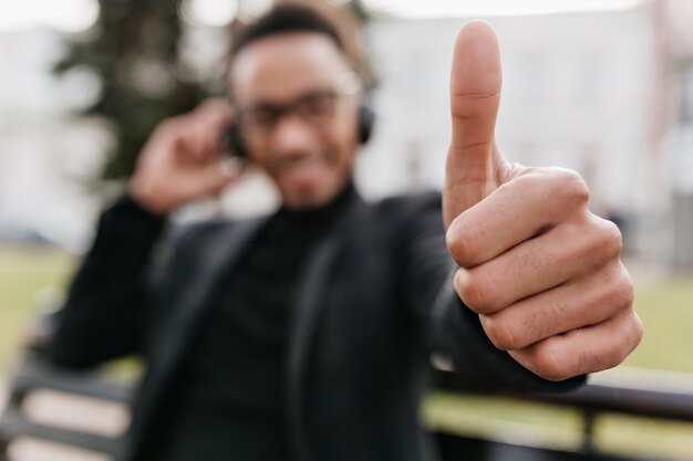 Sfocatura ritratto di africano ragazzo sorridente con la sua mano a fuoco. uomo di colore rilassato in abito elegante seduto sulla panchina e mostrando il pollice in su.