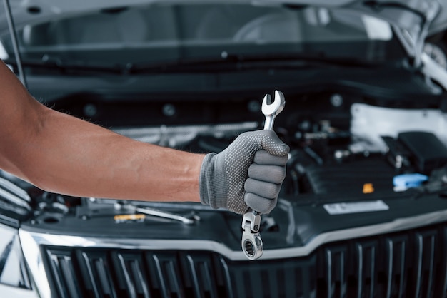 Blurred background. Man's hand in glove holds wrench in front of broken automobile