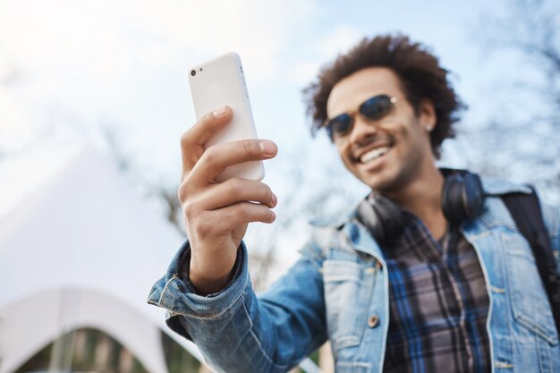 ガジェットで笑って公園でスマートフォンでselfieを取っている間デニムの服とメガネを着て、アフロの髪型と毛で興奮した浅黒い肌の男のぼやけた屋外のポートレート。