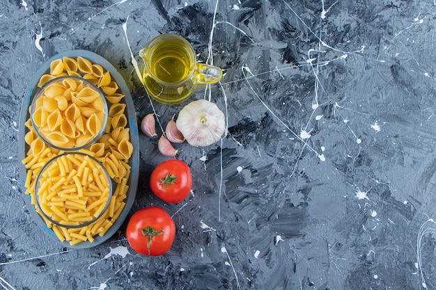 A board of two types of raw macaroni with vegetables and oil on a marble background .