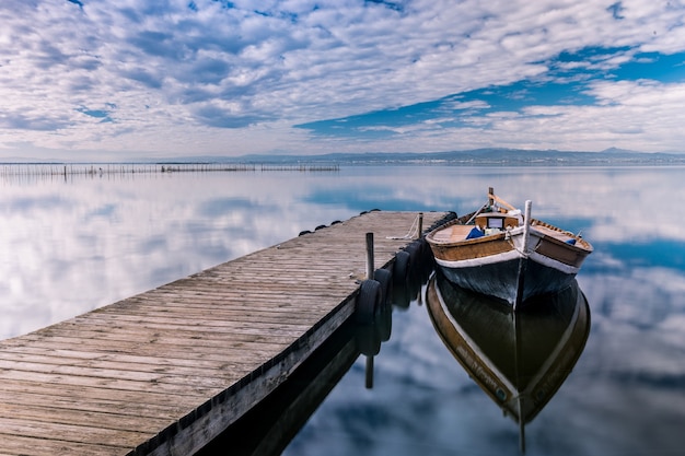 Foto gratuita barca parcheggiata vicino al molo di legno con riflessi nel mare sotto il cielo nuvoloso