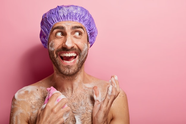 Free photo body care concept. happy guy takes shower, relaxes after stressed work, has wet skin with foam, rubs chest with sponge, looks joyfully aside, isolated on pink wall