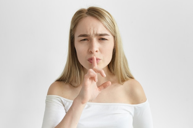 Body language. Attractive European girl in stylish top holding fore finger at her lips and frowning, asking to keep silent. Stylish young blonde female making hush gesture, saying Hush, be quiet