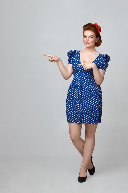 Body language. Attractive fashionable young woman wearing bright make up and vintage outfit standing isolated , pointing fingers, indicating copyspace wall for your text or advertisement