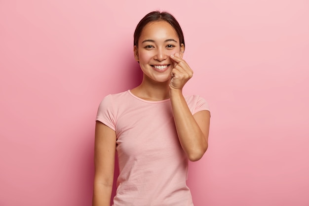 Body language concept. Happy Asian woman makes korean hand sign, expresses love, makes like gesture, smiles gently , dressed in casual wear, isolated on pink wall. Monochrome.