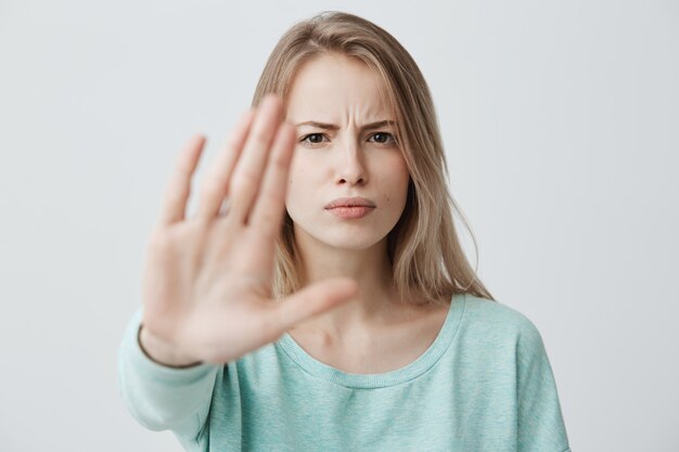 Body language. Disgusted stressed out angry blonde young female with straight fair hair posing against wall, keeping hand in stop gesture, trying to defend herself as if saying: Stay away from