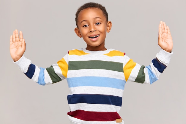 Body language.  handsome adorable Afro American boy in striped pullover keeping hands wide apart as if holding something very large in size, measuring, having excited expression