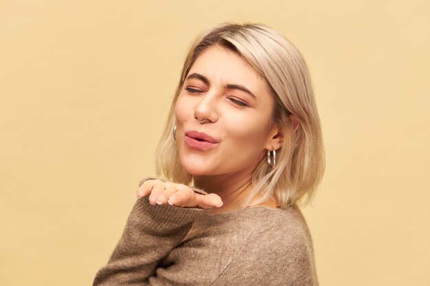 Body language. Horizontal shot of charming amazing young European female dressed in cozy cashmere sweater having playful look, flirting with you, pouting lips and keeping hand open, blowing kiss